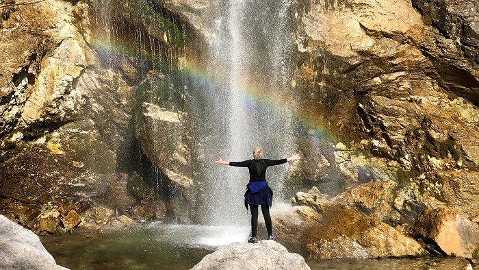 Ein Mädchen steht unter einem Wasserfall, auf dem sich ein Regenbogen gebildet hat