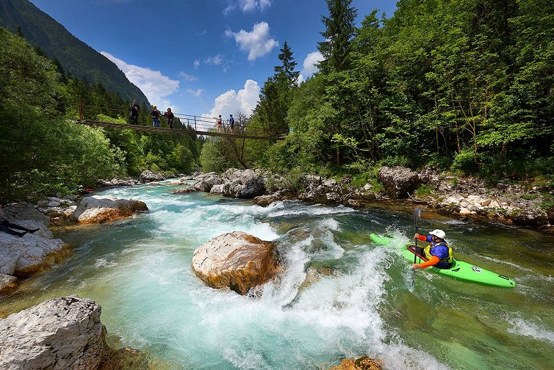 The kayaker is descending the Soča River and will go under the hanging bridge, where tourists are watching him.