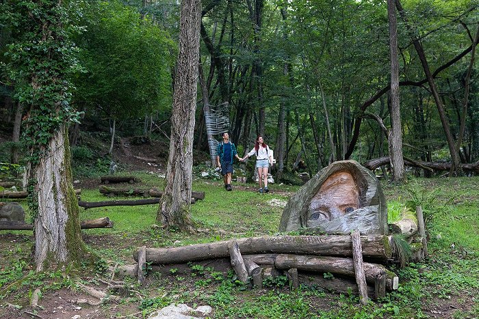 Two visitors walk through the park, where they are viewed by an interesting wooden sculpture