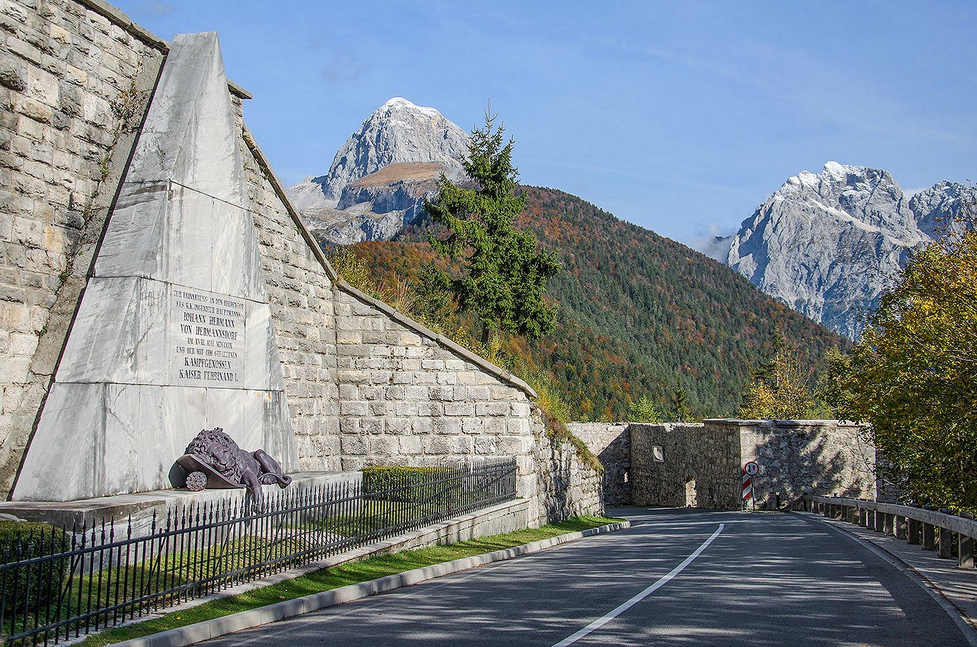 Spomenik s kipom ranjenega leva ob cesti proti Bovcu, v ozadju gora Mangart