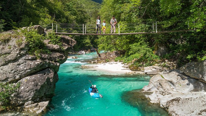 Der Kajakfahrer steigt unter einer Hängebrücke den Fluss Soča hinab, wo er von einer Familie von Wanderern beobachtet wird.