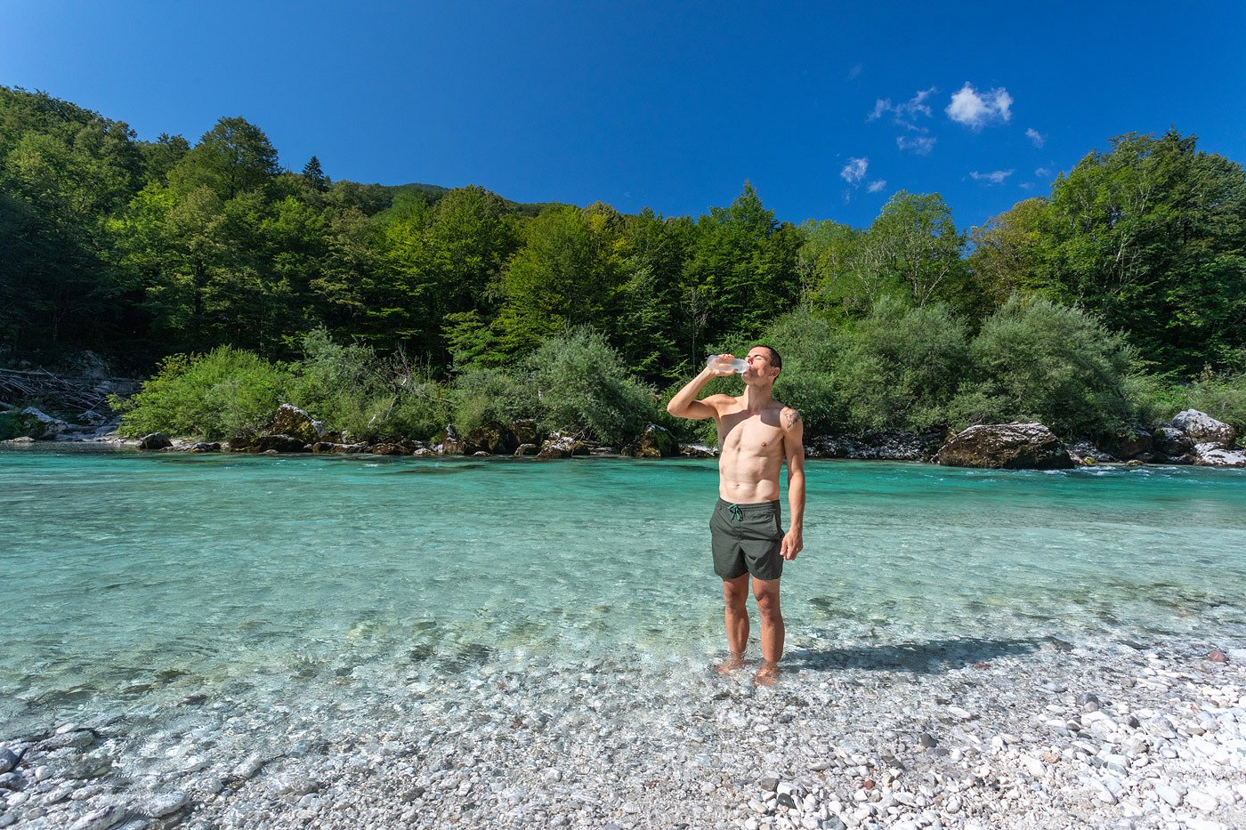 A tourist in a bathing suit stands in the Soča River and drinks water from a bottle