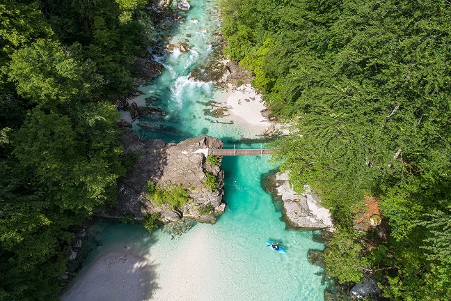 A bird's eye view of the Soča River, which is crossed by a hanging bridge. A kayaker paddles on the Soča.