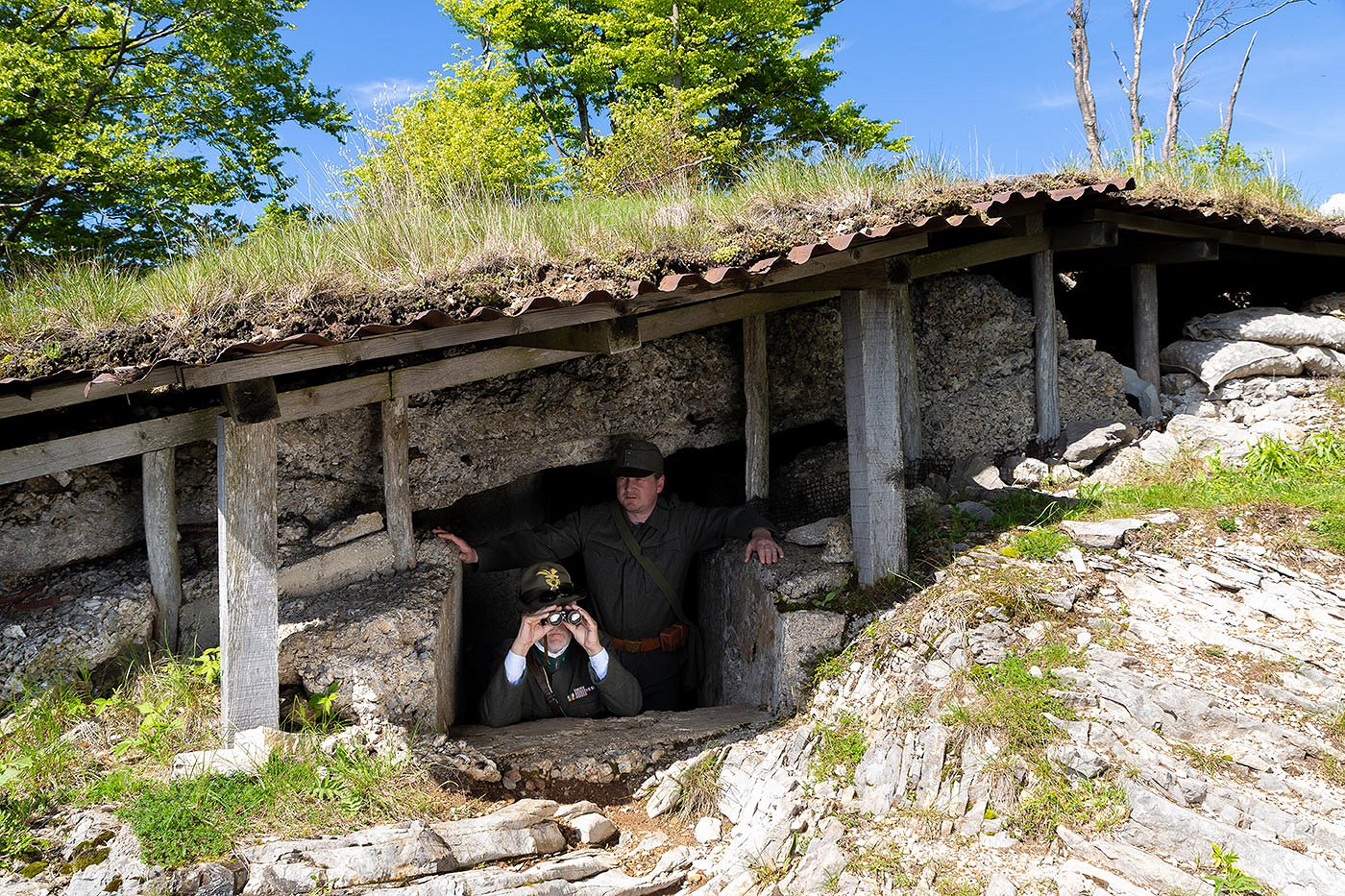 Two soldiers in World War I uniforms observe the surroundings from a ditch with binoculars