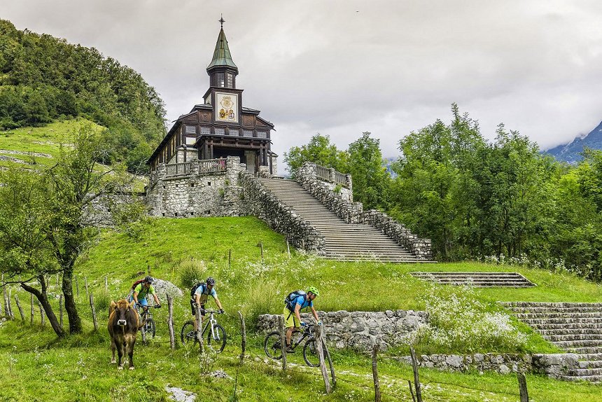 Trije gorski kolesarji se spuščajo po potki pod leseno cerkvijo v Javorci kjer pase tudi krava.