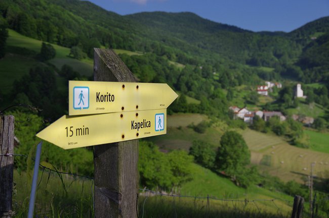 Un cartello giallo su un palo di legno che mostra la direzione. Sullo sfondo un villaggio.