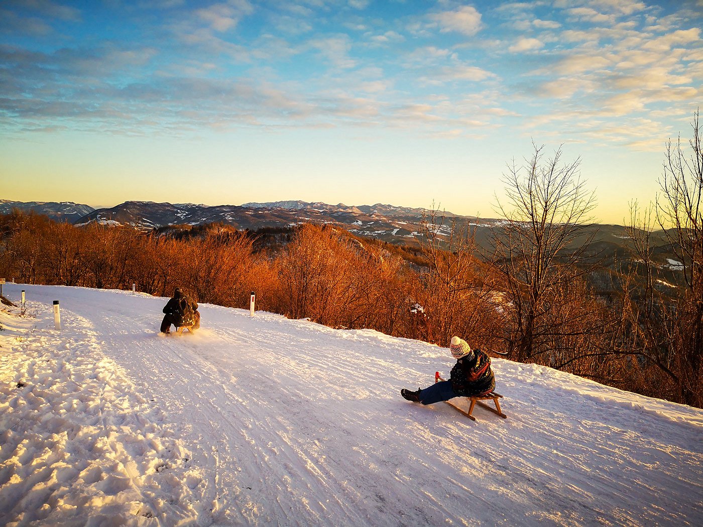 Two people are sledding down a snowy road at a golden sunset.