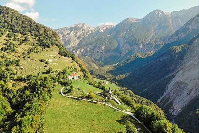Auf der linken Seite des Bildes befinden sich auf einem sonnigen, grasbewachsenen Hang die Gedächtniskirche des Heiligen Geistes in Javorca und ein nahegelegenes Gehöft. Die Wiese ist von einem gelb werdenden Laubwald umgeben. Rechts öffnet sich der Blick auf einen sonnigen Bergrücken, der von Wald bedeckt ist, während der untere Teil einen schattigen, sandigen Hang zeigt, der von einem Erdrutsch betroffen ist.